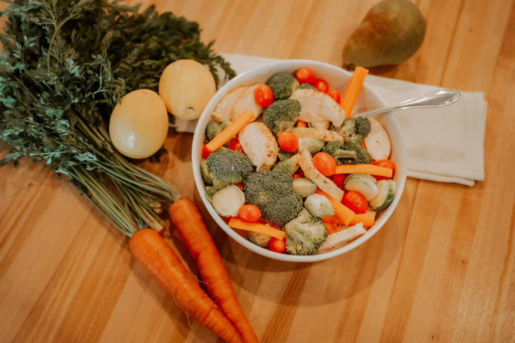A plate full of fresh vegetables like cherry tomatoes, carrot sticks, broccoli florets and roasted chicken. Carrots and lemons and a pear off to the side.