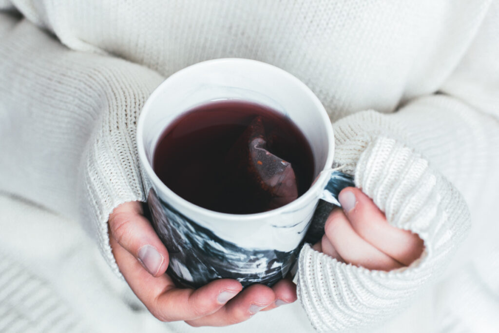 Woman wearing a cozy sweater holding a mug of hot tea with a tea bag dangling inside.