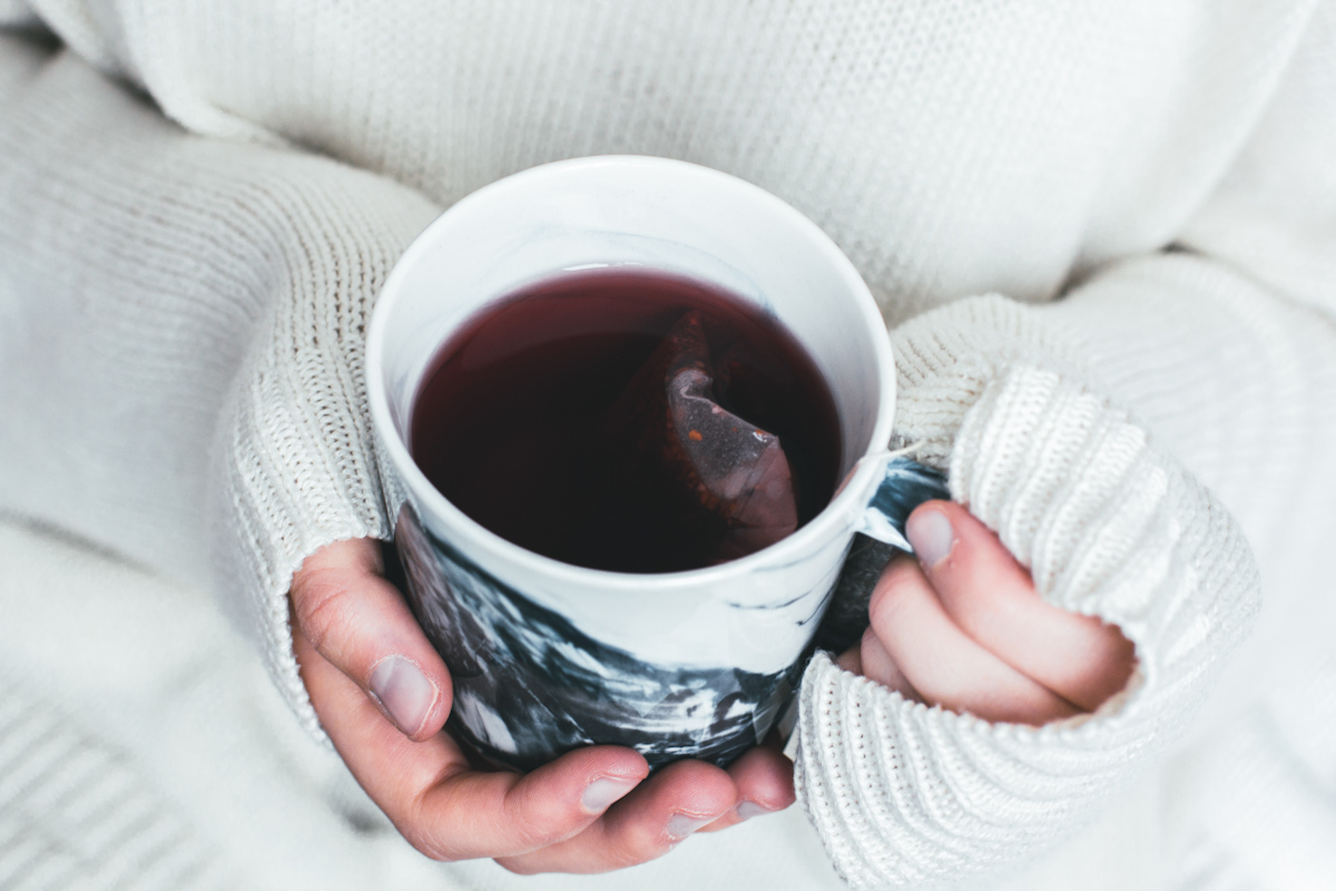 Woman wearing a cozy sweater holding a mug of hot tea with a tea bag dangling inside.