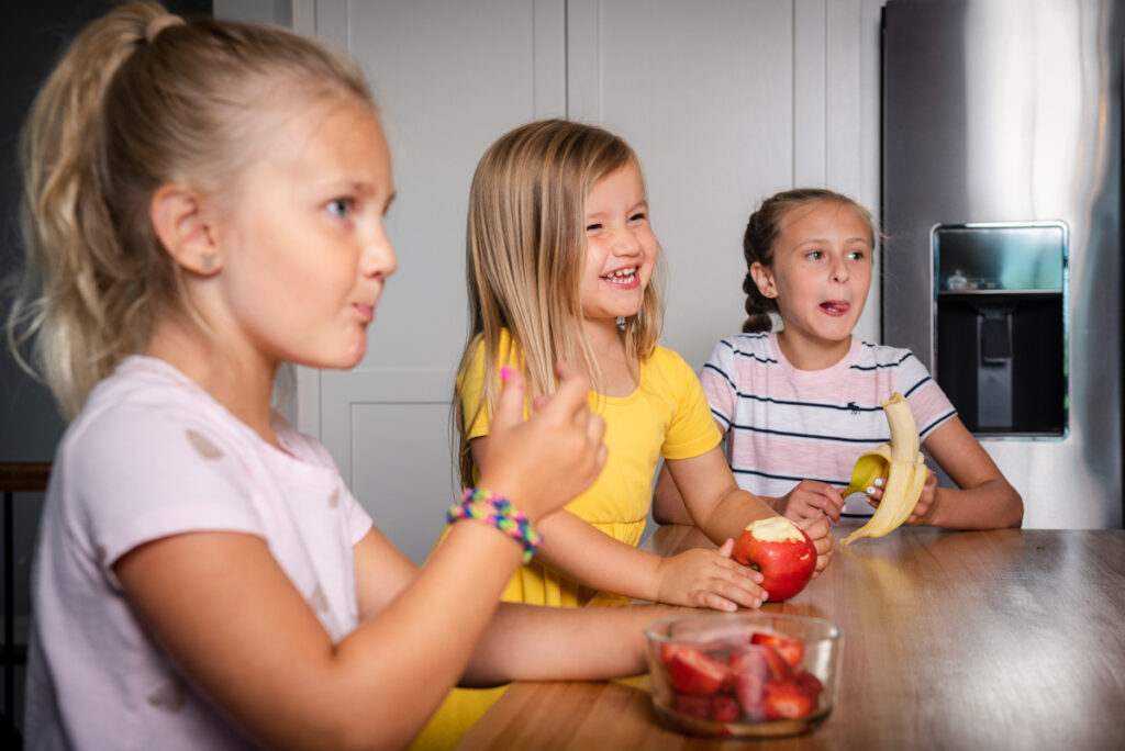 Three little girls snacking on apples, strawberries, and bananas