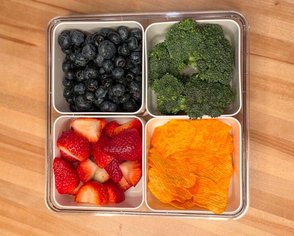 A four section snack container with strawberries, blueberries, broccoli florets, and healthier potato chips.