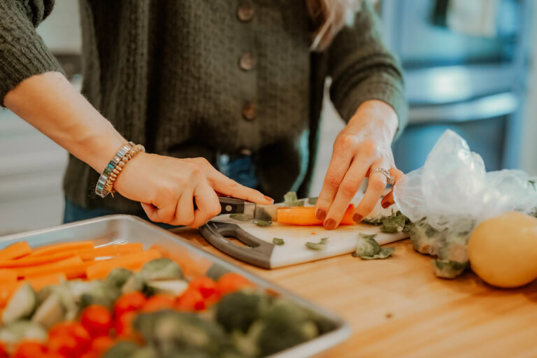 A woman cutting carrots with a baking sheet full of chopped broccoli, cherry tomatoes, and chopped brussels sprouts.