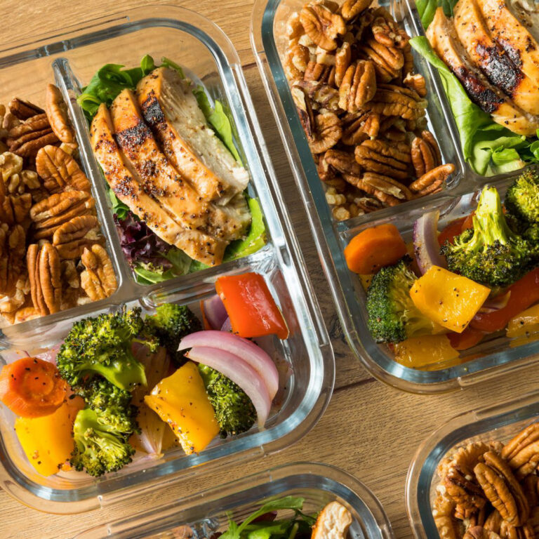 4 meal prep clear glass containers with grilled chicken, steamed broccoli, peppers, carrots, and onions and sliced pecans.