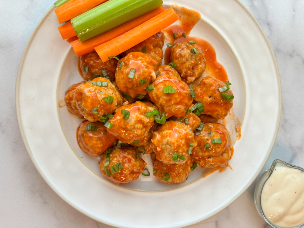 A plate of colorful, buffalo chicken meatballs topped with green onions with a side of ranch and a side of carrots and celery