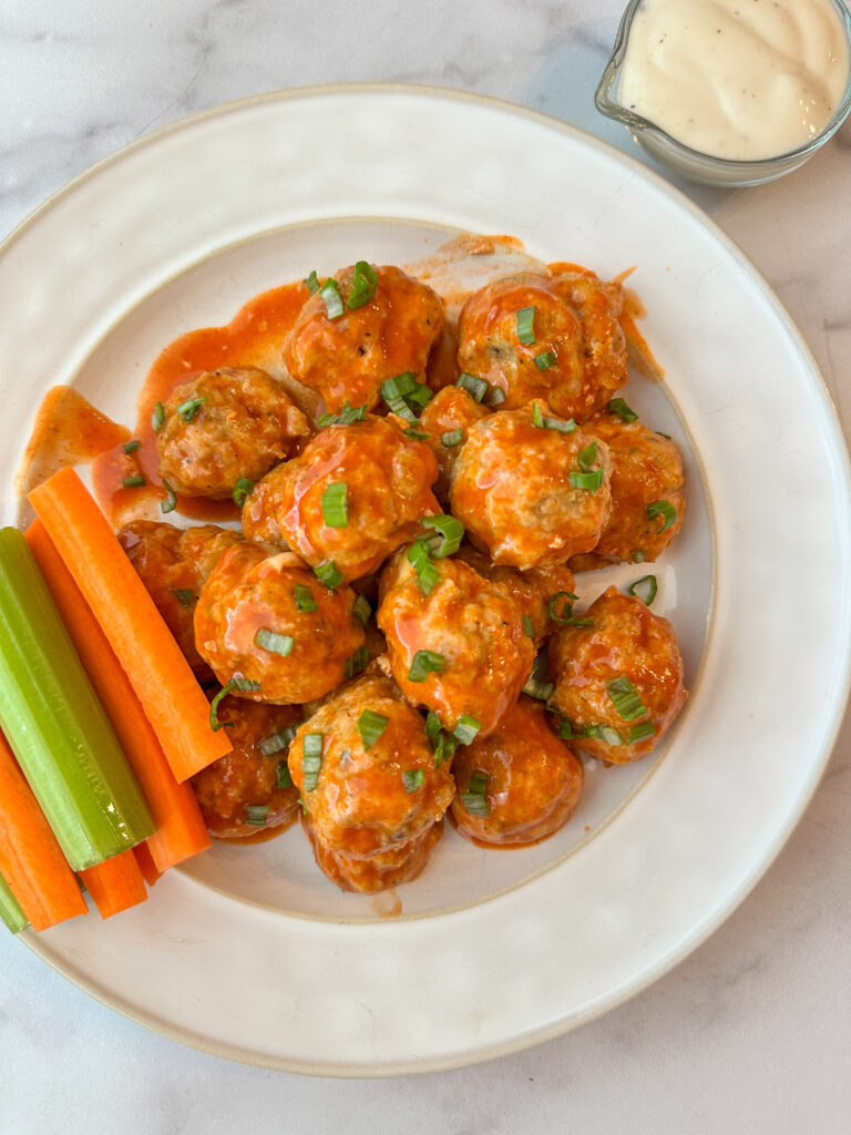 A plate of colorful, buffalo chicken meatballs topped with green onions with a side of ranch and a side of carrots and celery