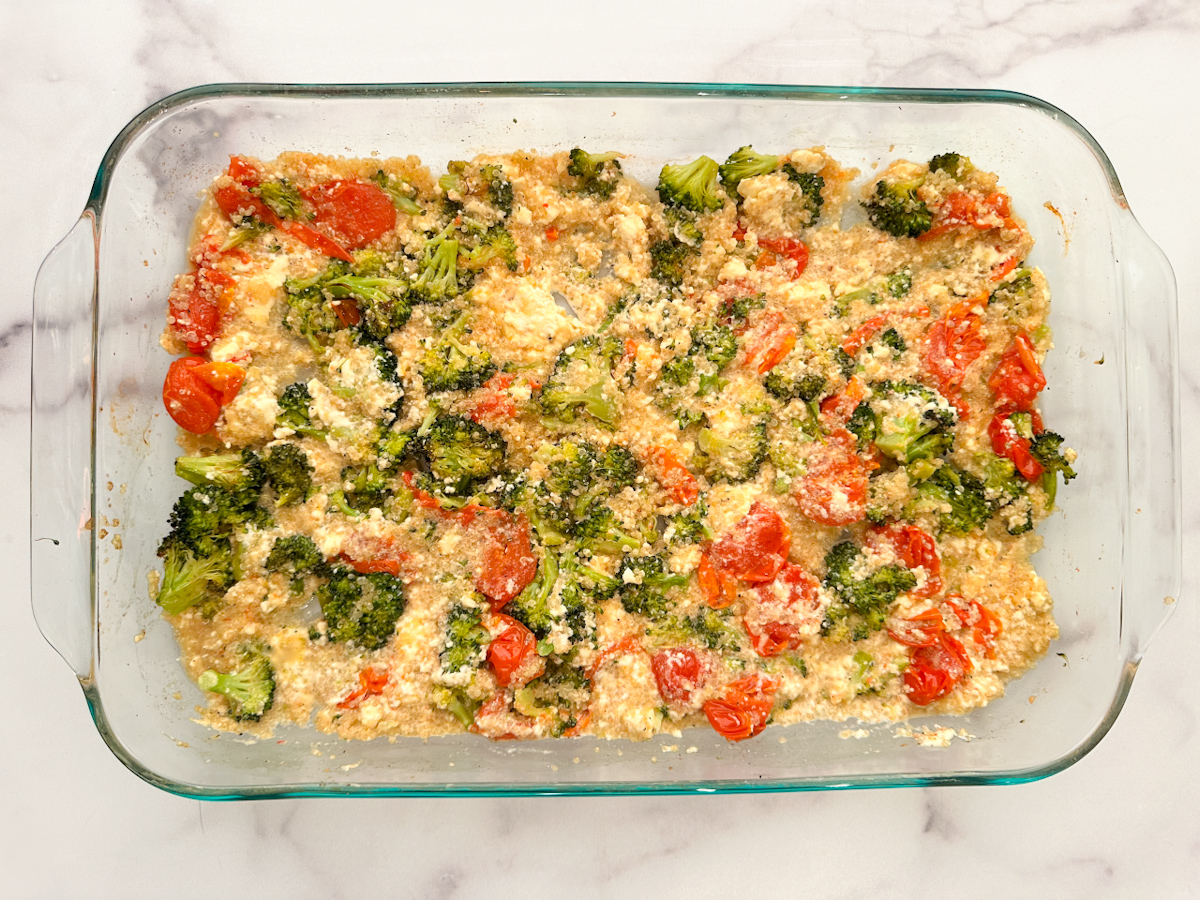 Baked Chicken Quinoa Veggie and Feta Bake in a large clear baking dish with chunks of chicken, broccoli, tomatoes, quinoa and melted feta cheese.