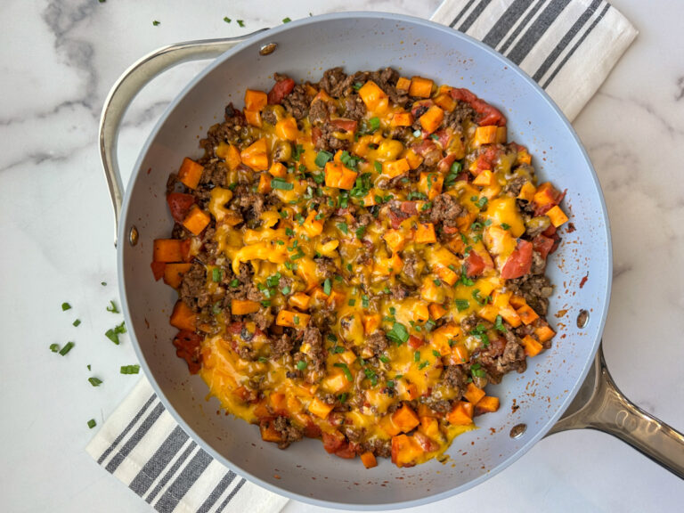 Non-stick skillet filled with cooked ground beef, orange sweet potatoes, tomatoes, green onion garnish.