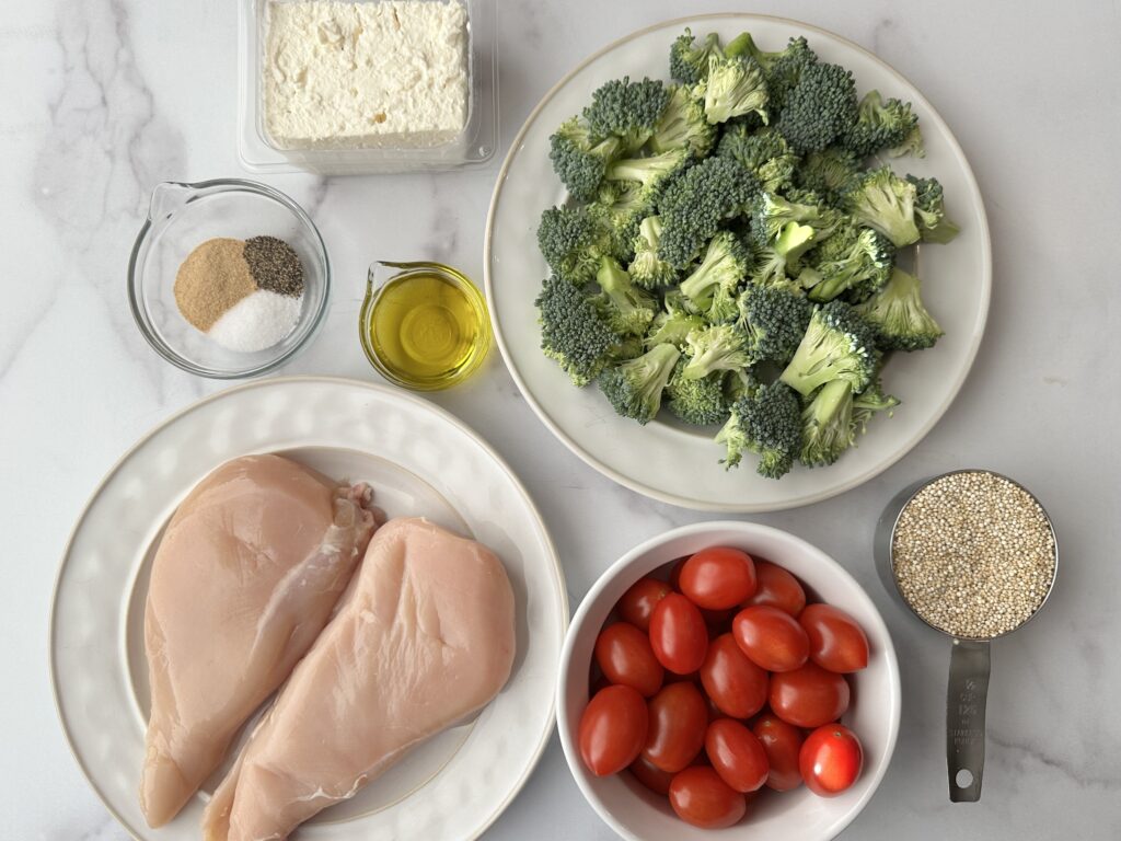 white countertop with white dishes full of green broccoli, red tomatoes, two boneless, skinless chicken breasts, a bowl of seasonings, a small dish of olive oil, a block of feta cheese, and a measuring cup with quinoa.