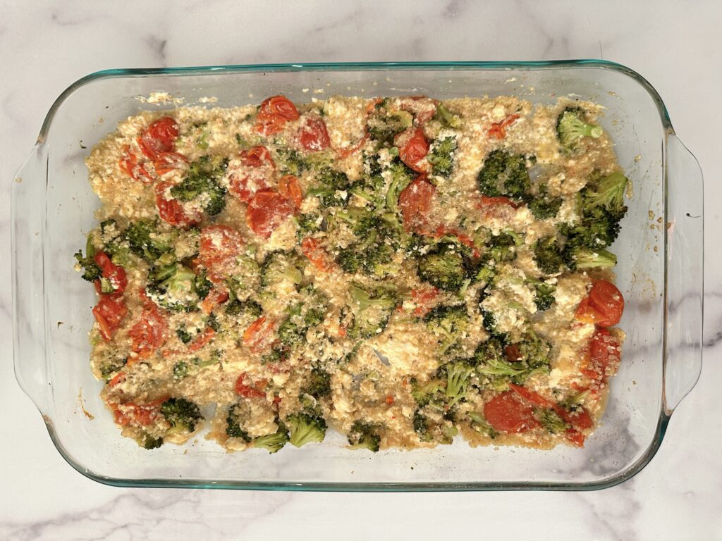 A clear casserole dish with baked broccoli, tomatoes, quinoa, and feta cheese mixed up together.