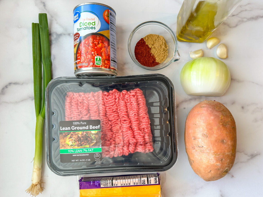 A white background with a package of raw ground beef, one sweet potato, a white onion, three garlic cloves, olive oil bottle, spices, can of diced tomatoes, a block of cheddar cheese, and a green onion.