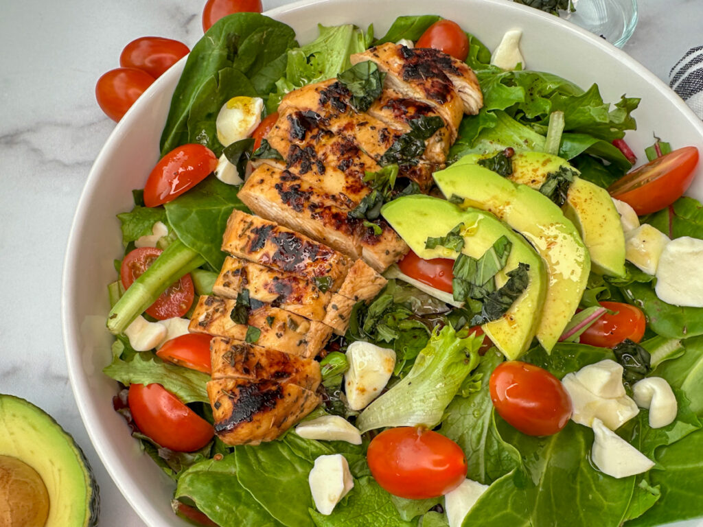White bowl full of mixed greens, grilled marinaded chicken, sliced tomatoes, fresh mozzarella, and sliced avocado.