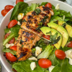 White bowl full of mixed greens, grilled marinaded chicken, sliced tomatoes, fresh mozzarella, and sliced avocado.