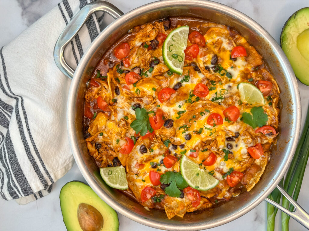 A cheesy chicken enchilada skillet in a stainless steel skillet with tomatoes, black beans, chicken, avocado, and tortilla quarters