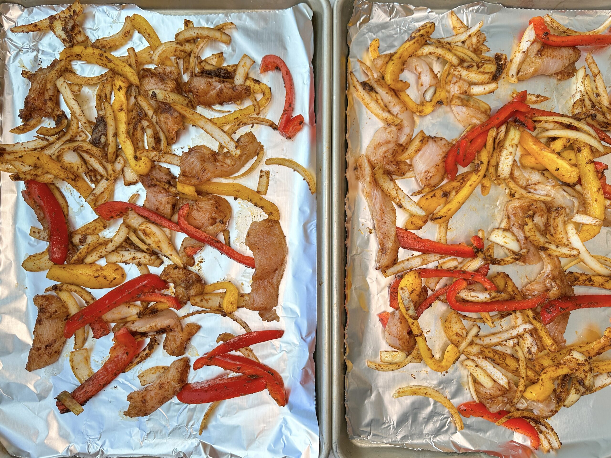 Sheet pan fajita ingredients spread out over two baking sheets: Sliced chicken, red and yellow bell peppers, onions and seasonings.