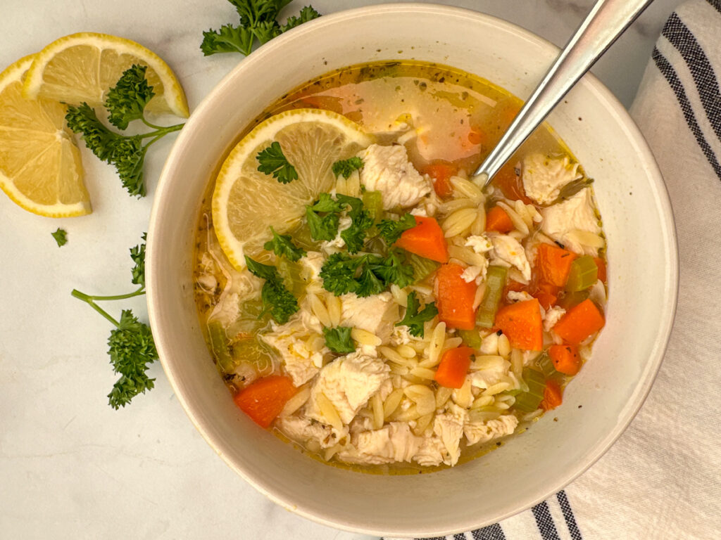 White bowl of soup with chopped chicken, orzo, celery, carrots, parsley, and lemon