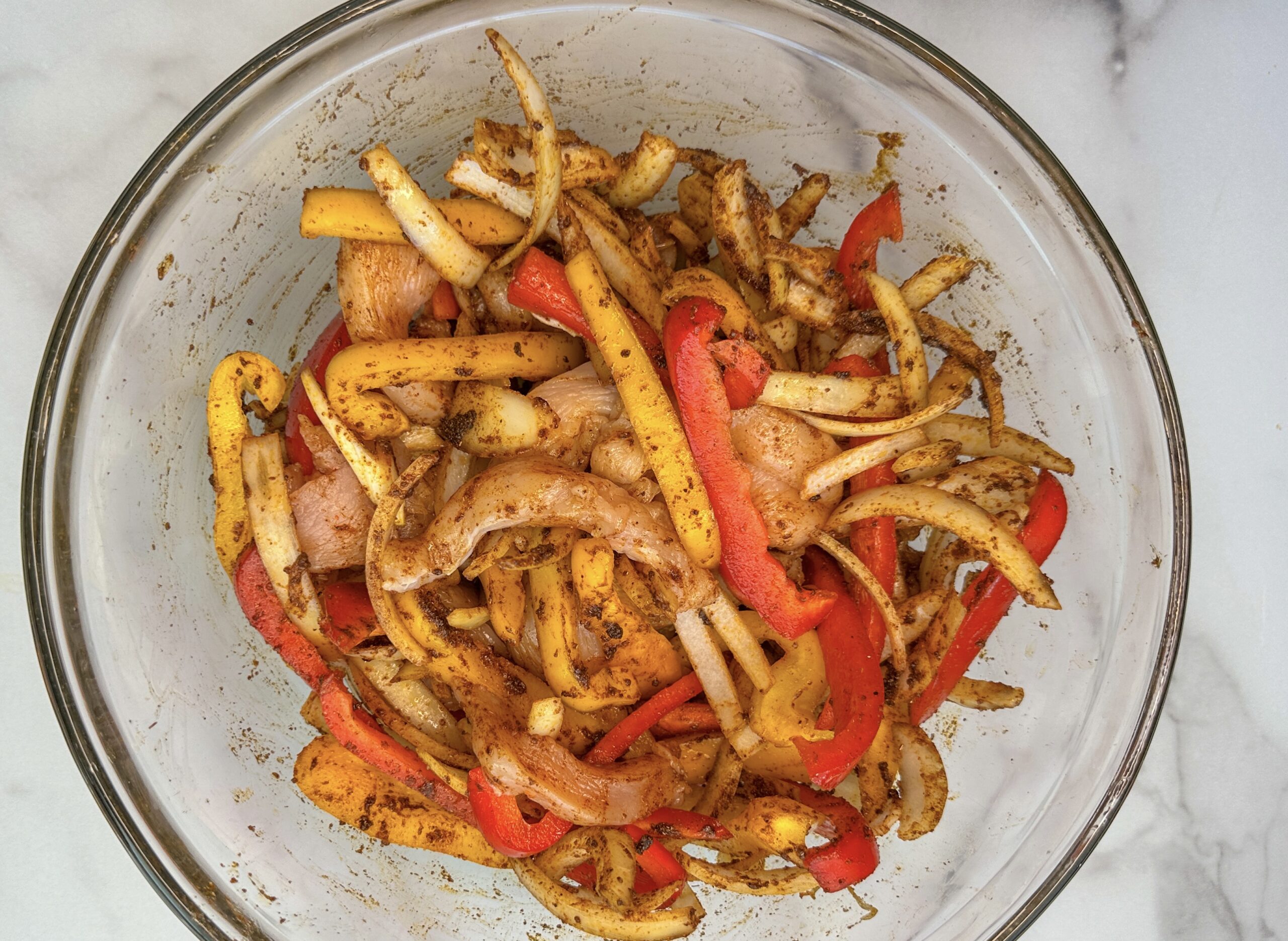 sheet pan fajita ingredients: chicken breast strips, sliced red and yellow bell peppers, and sliced onion with seasonings all mixed up into a mixing bowl.