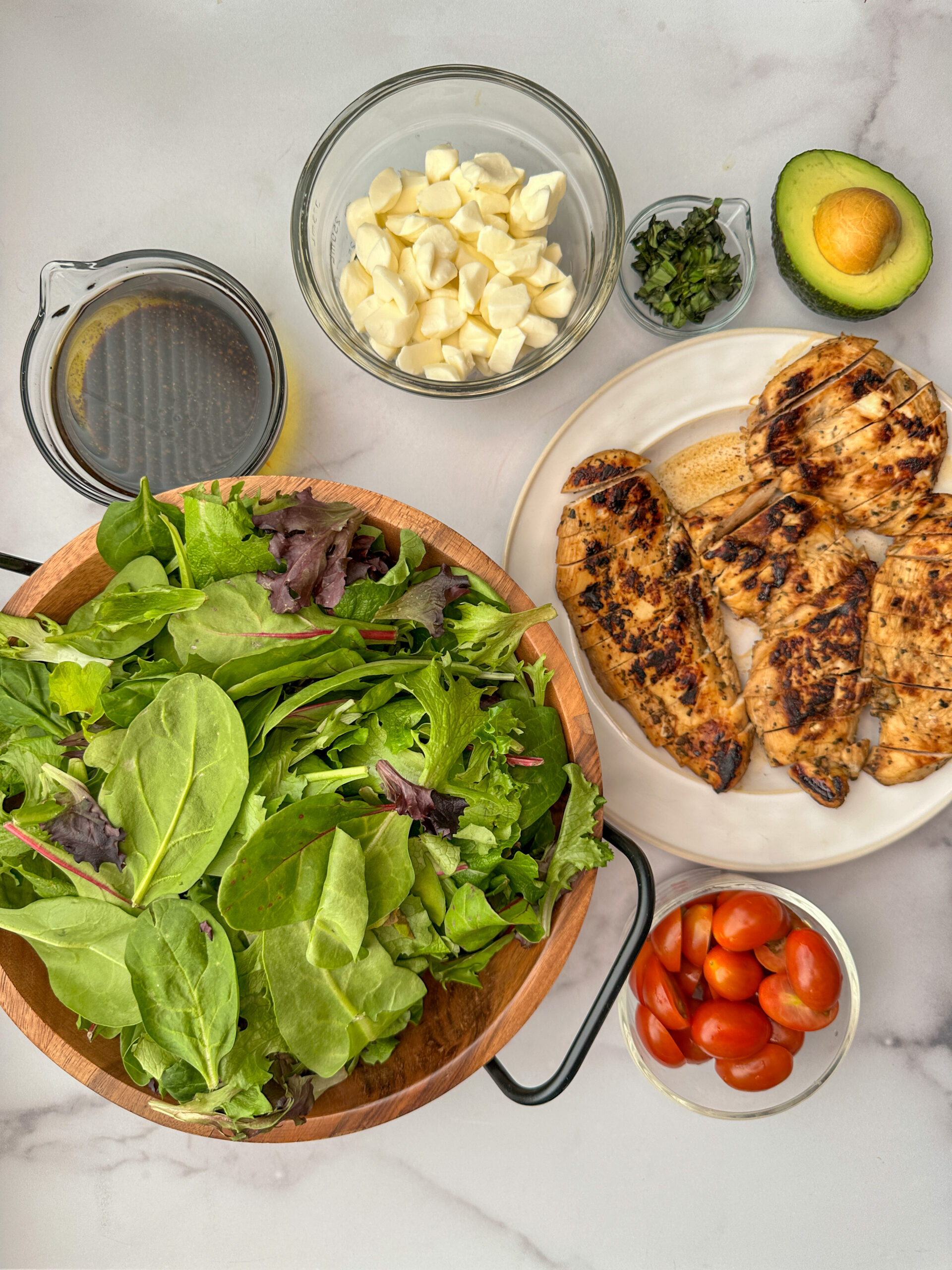 Balsamic chicken caprese salad ingredients: mixed greens, marinade, mozzarella, basil, avocado, chicken breasts, and tomatoes.