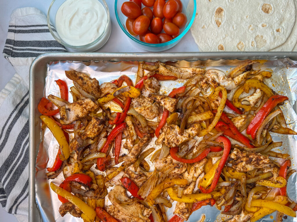 Sheet pan with roasted chicken and sliced red and yellow bell peppers, sliced translucent onions and a bowl of sour cream, tomatoes, and tortilla shells.