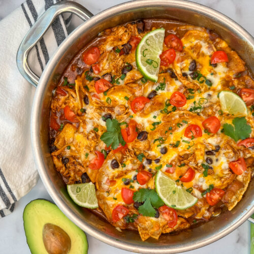 A cheesy chicken enchilada skillet in a stainless steel skillet with tomatoes, black beans, chicken, avocado, and tortilla quarters