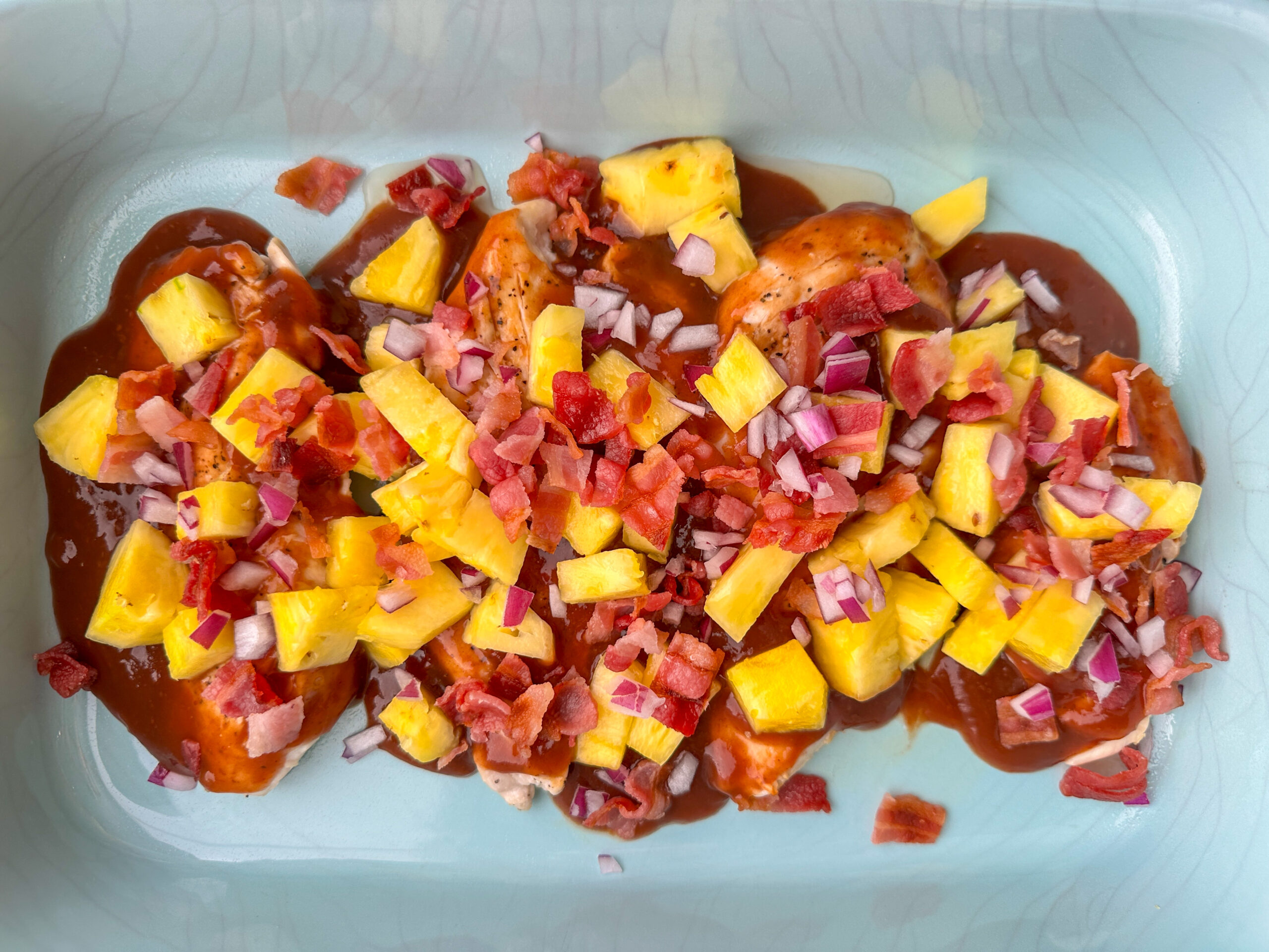 four grilled chicken breasts topped with bbq sauce, bacon, and pineapple in a large baking dish.