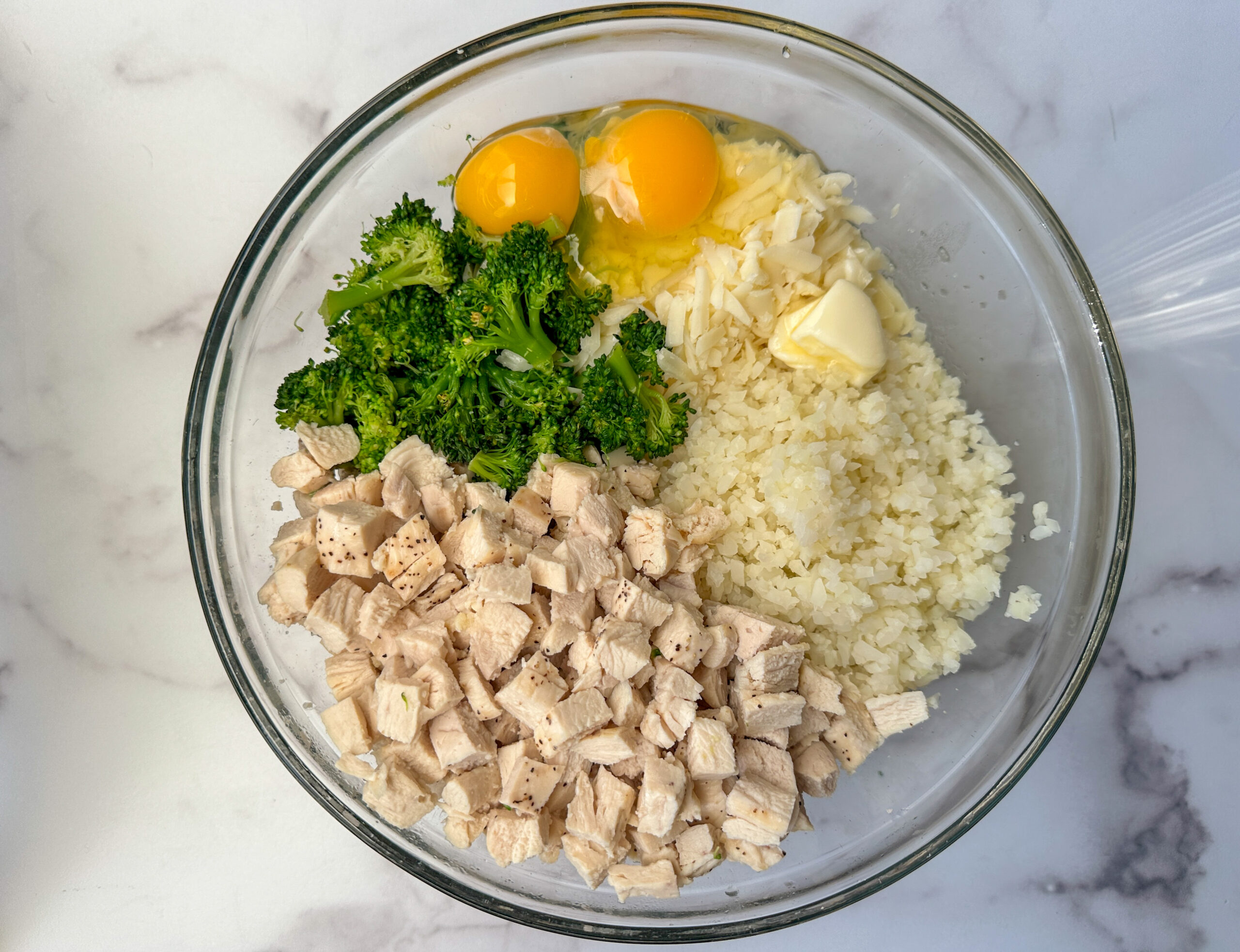 Broccoli Cauliflower Ingredients mixed into a large bowl: chicken, broccoli, 2 eggs, butter, shredded cheese, and cauliflower rice.