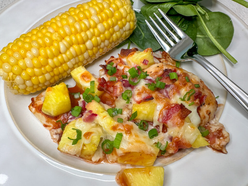 Chicken breast smothered in pineapple, bbq sauce, cheese, and green onions with corn on the cob and spinach salad on the plate.
