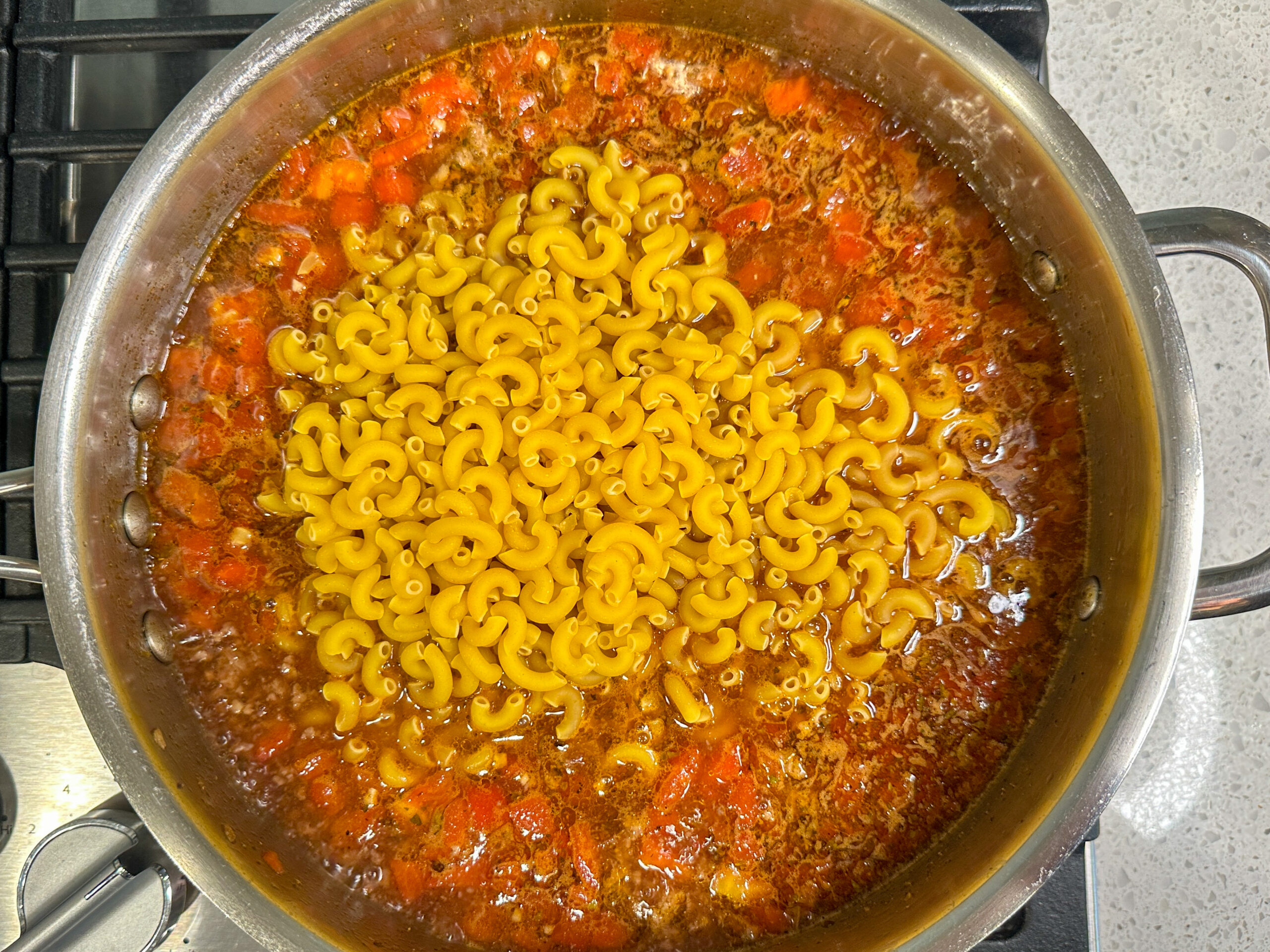 Skillet with beef broth, diced tomatoes, and uncooked pasta noodles on top.