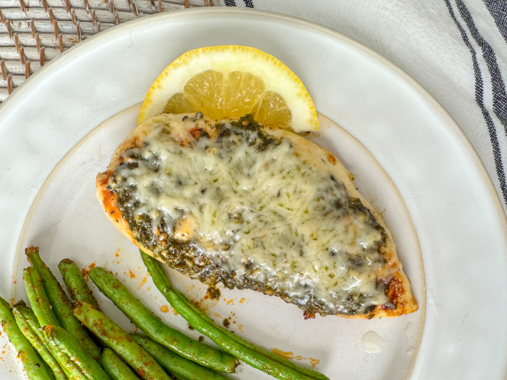 Grilled lemon pesto chicken on a white plate with a grilled chicken breast topped with basil pesto, melted parmesan cheese and a lemon wedge