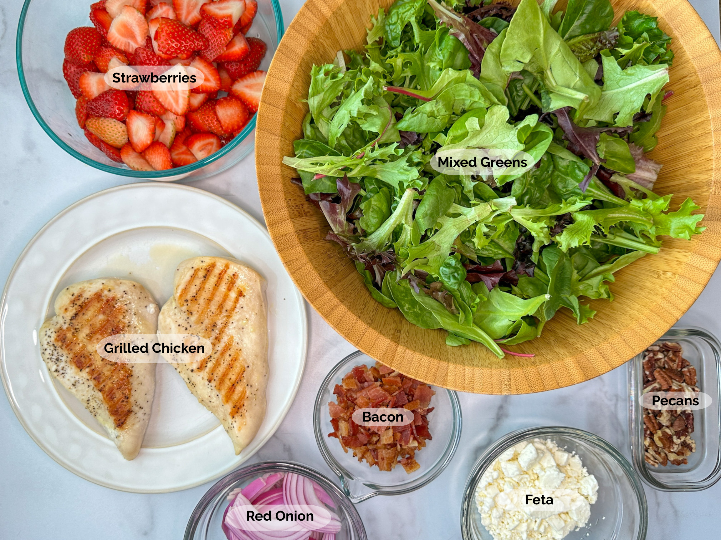 Strawberry Fields Salad ingredients: halved strawberries, a bowl of mixed greens, 2 grilled chicken breasts, chopped cooked bacon, sliced red onions, crumbled feta, and chopped pecans.