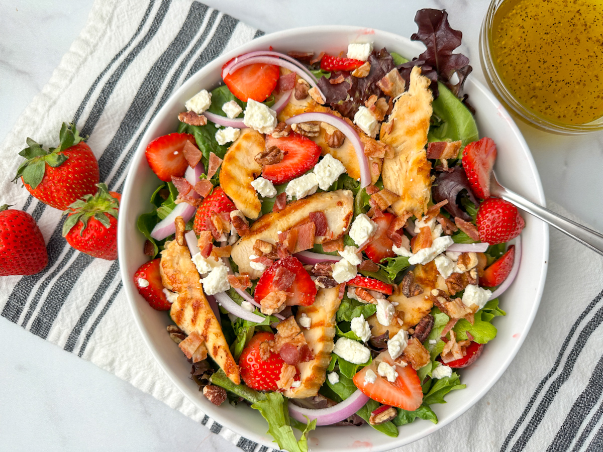 Strawberry Fields Salad in a plate bowl with mixed greens, strawberries, crumbled feta cheese, sliced red onion, sliced grilled chicken, bacon pieces, and the salad dressing on the side.