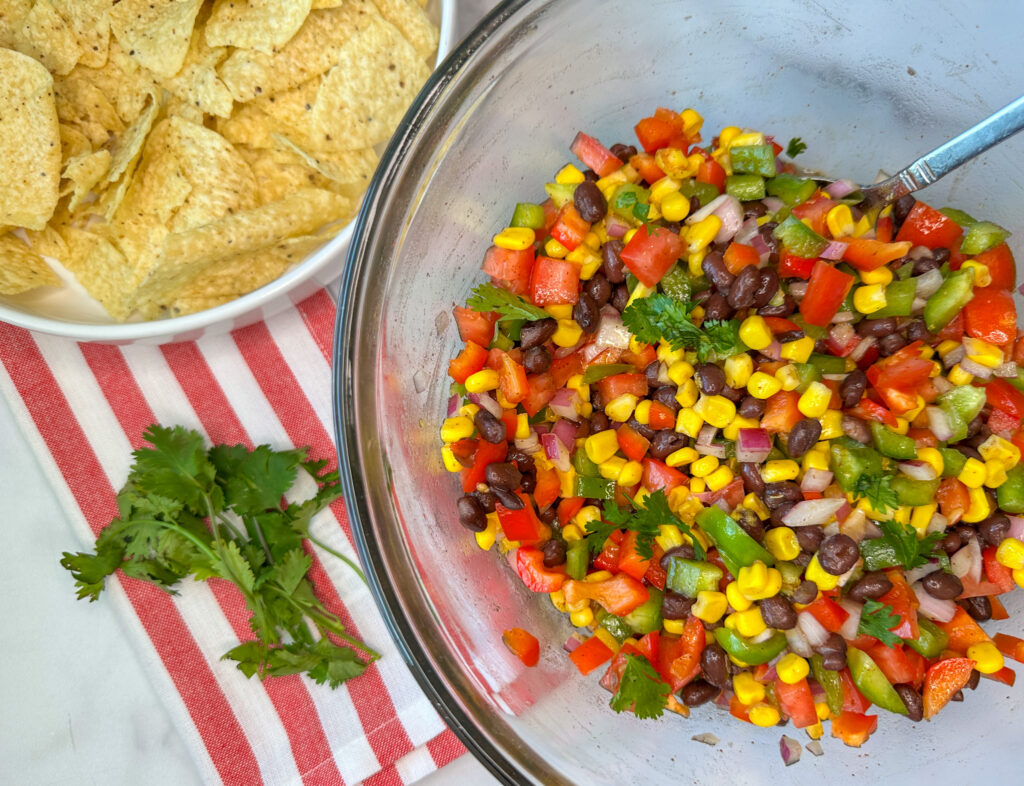 a glass bowl full of cowboy caviar which has chopped green and red bell peppers, chopped red onion, chopped roma tomatoes, black beans, and corn with cilantro mixed in.