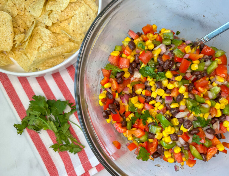 a glass bowl full of cowboy caviar which has chopped green and red bell peppers, chopped red onion, chopped roma tomatoes, black beans, and corn with cilantro mixed in.