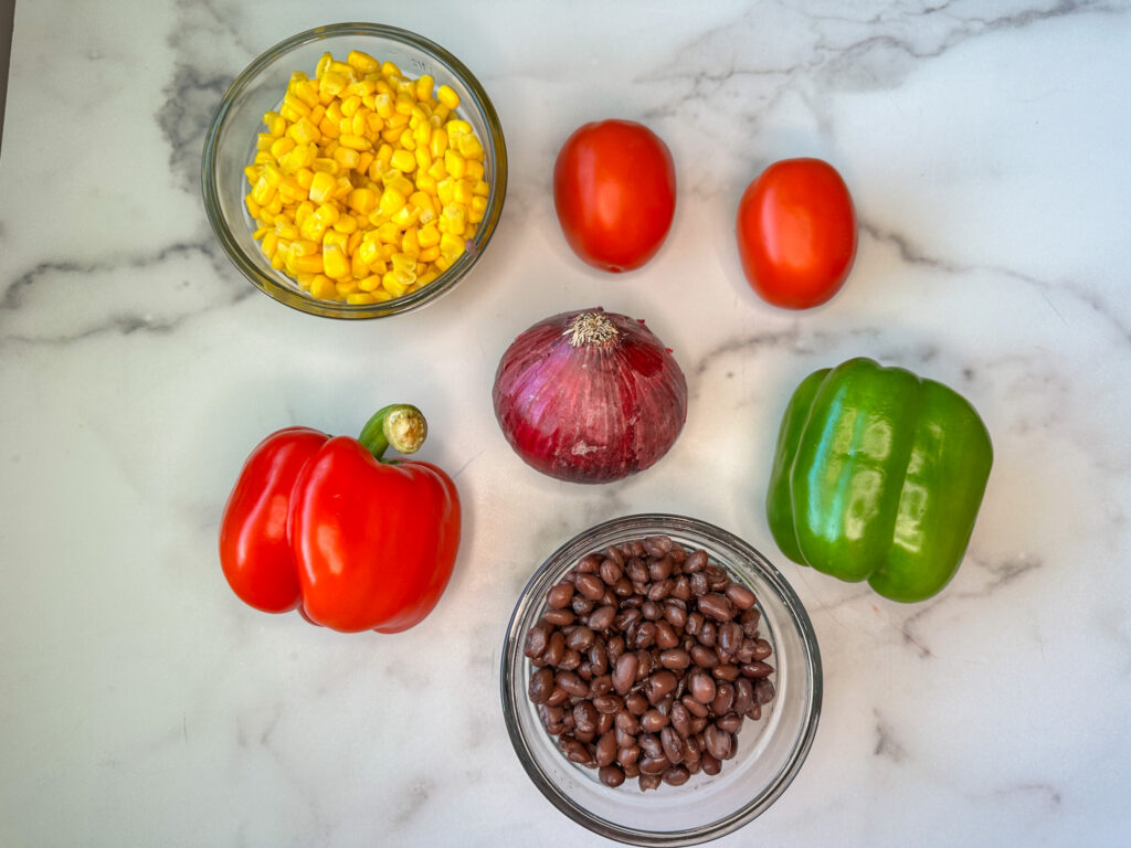 Healthy cowboy caviar ingredients - red and green bell peppers, two Roma tomatoes, a bowl of black beans, a bowl of corn, and a red onion