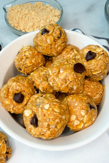 lower calorie protein balls in a white bowl. Several protein balls in the bowl with a few laying off to the side. A side dish of oats and chocolate chips are to the side.