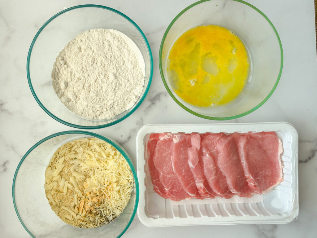 Crispy parmesan pork chops ingredients: raw thin pork chops, a bowl of flour, a bowl of a beaten egg, and a bowl of panko bread crumbs, parmesan cheese, and seasonings.