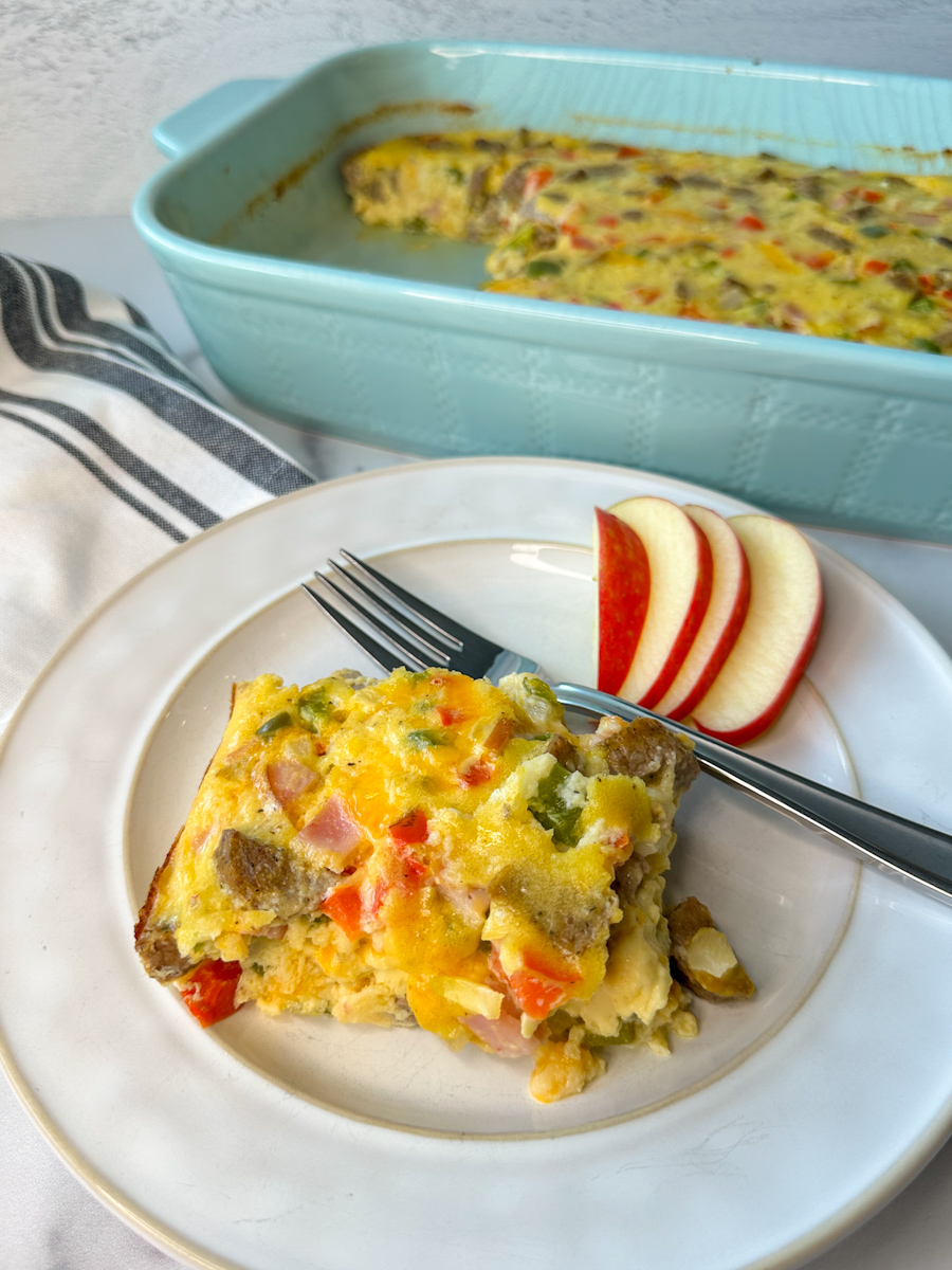 Egg and cottage cheese bake with diced red and green bell peppers and onions, chunks of turkey sausage and Canadian bacon and a side of sliced apples on a plate.
