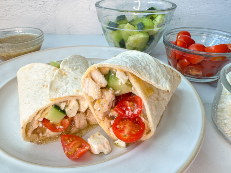 Grilled Greek Chicken Wrap with diced, cooked chicken, diced tomatoes and diced cucumbers wrapped up in a tortilla, cut in half, and sitting on a plate