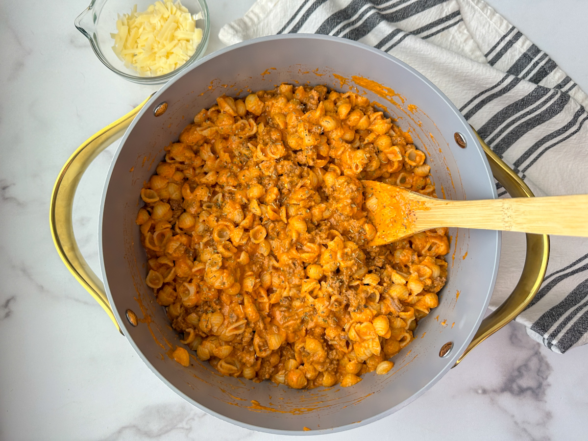 A large skillet with cooked high protein pasta: small shell noodles with lean ground beef and an orange sauce consisting of cottage cheese and marinara. A small side of shredded parmesan is on the side.