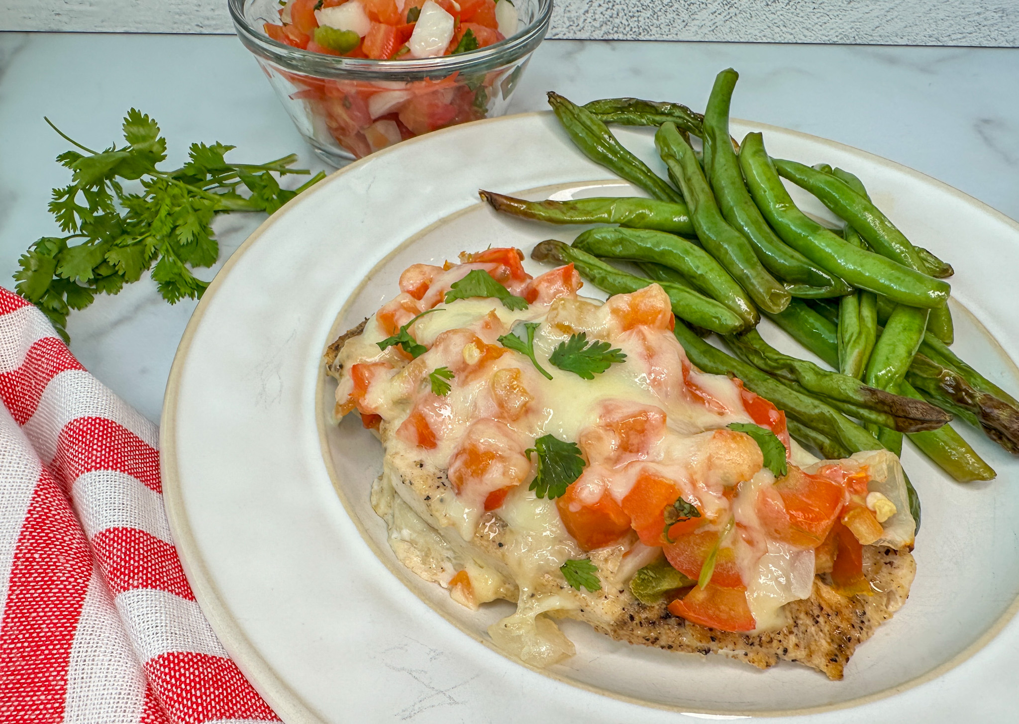 Baked Salsa Fresca chicken on a plate with a side of green beans. The chicken breast is topped wtih pico de gallo and melty Monterey jack cheese.