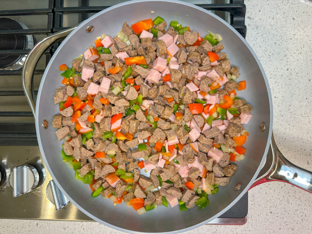 A large skillet with Canadian bacon, chopped sausage, red and green bell peppers and onions. 