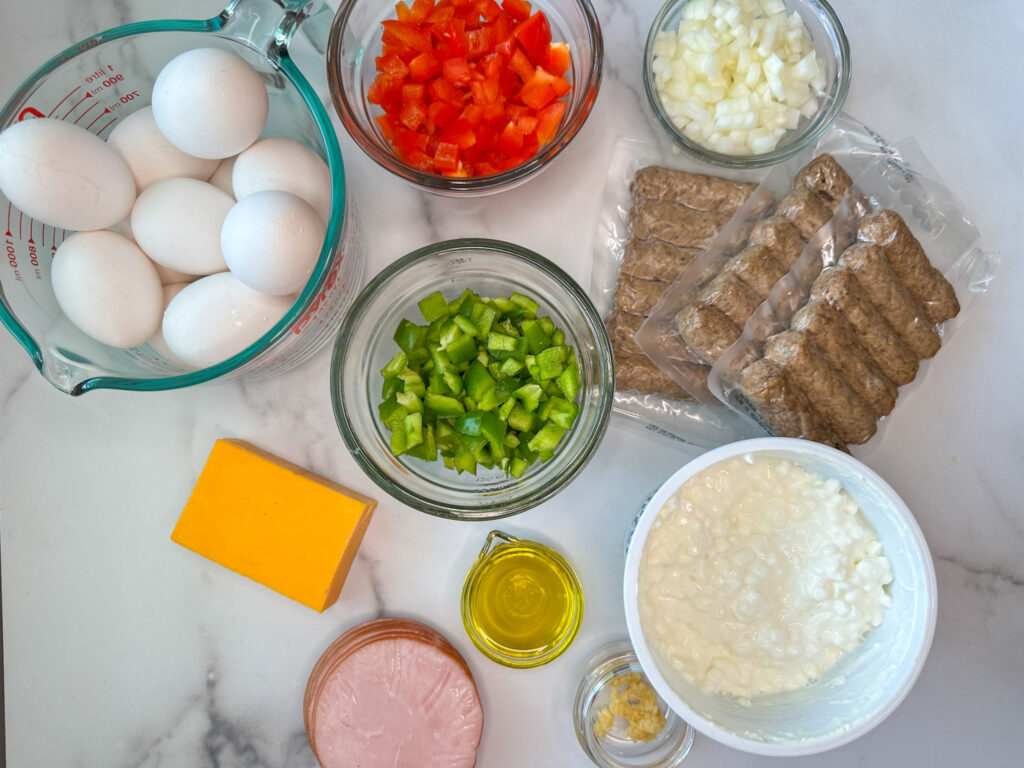 Egg and cottage cheese bake ingredients: a bowl of 12 eggs, a bowl of chopped red bell pepper, a bowl of chopped green bell pepper, a bowl of chopped sweet onion, 1 lb of sausage links, 16 oz of cottage cheese, 8 slices of Canadian bacon, 4 oz cheddar cheese block, minced garlic, and 1 tablespoon of olive oil. 