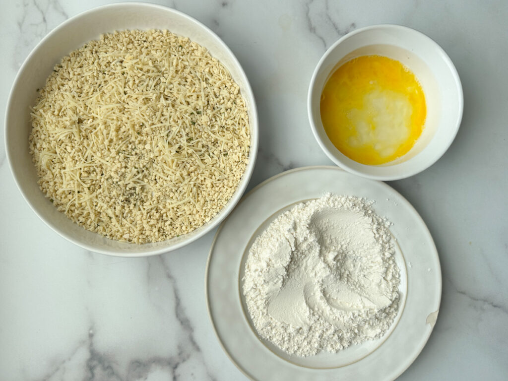 Large shallow dish with breading ingredients, white plate with flour on top, a white mixing bowl with a beaten egg.