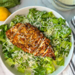 Crispy breaded chicken breast lying on a bed of romaine lettuce with shredded parmesan cheese with a lemon and dressing laying to the side