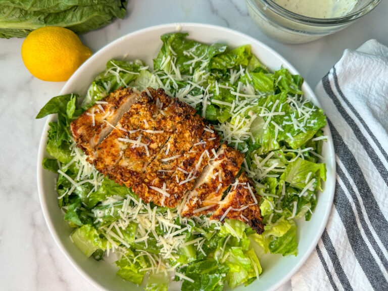 Crispy breaded chicken breast lying on a bed of romaine lettuce with shredded parmesan cheese with a lemon and dressing laying to the side