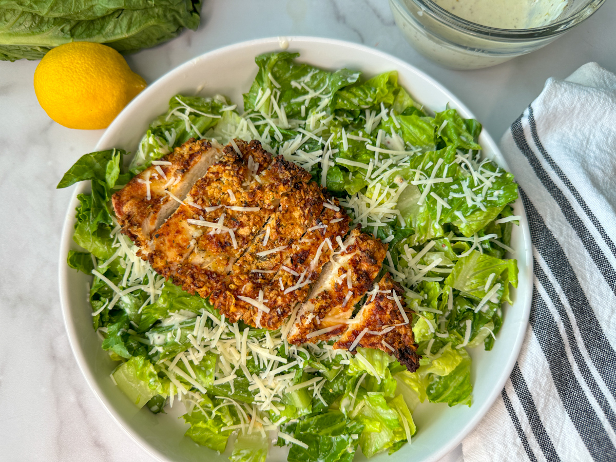 Crispy breaded chicken breast lying on a bed of romaine lettuce with shredded parmesan cheese with a lemon and dressing laying to the side
