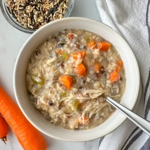 a bowl of chicken and wild rice soup with 2 carrots to the side and a small bowl of wild rice close by.