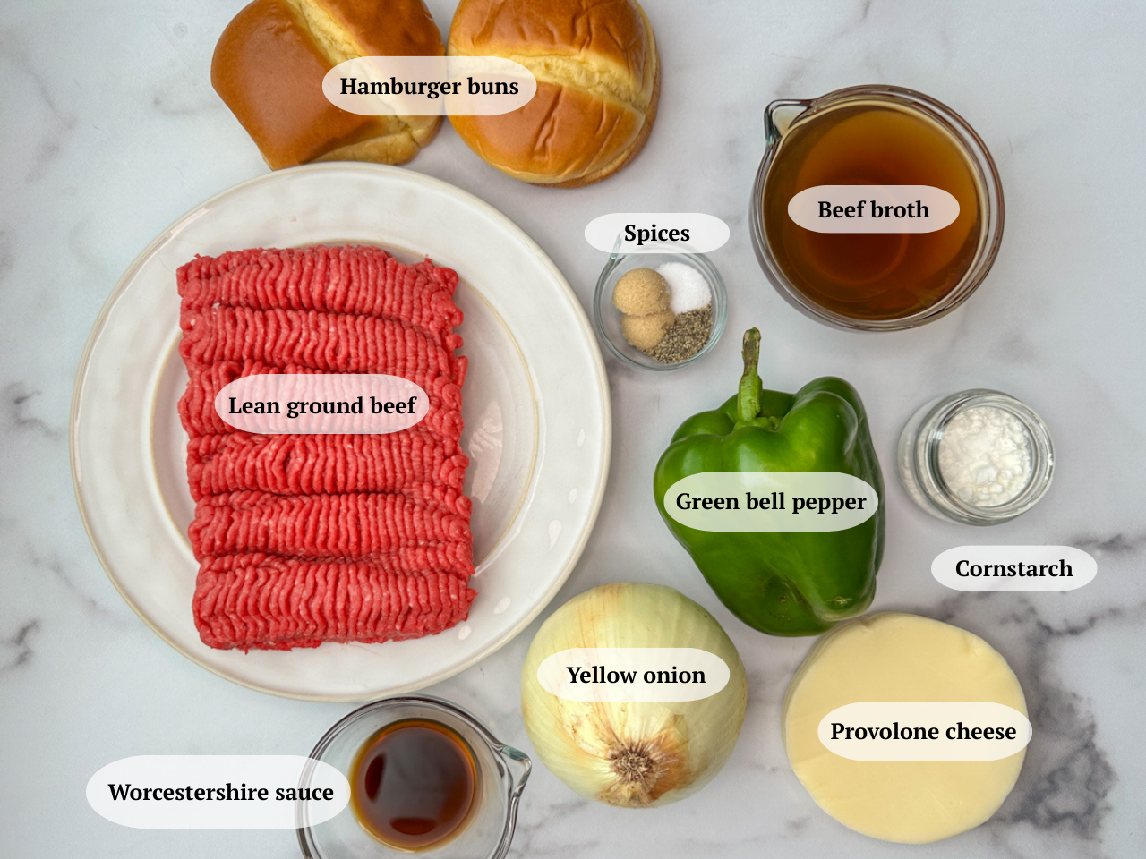 Philly cheesesteak sloppy joes ingredients - lean ground beef, 2 hamburger buns, a bowl of beef broth, a jar of spices, a green bell pepper, a yellow onion, a jar of cornstarch, provolone cheese slices, a small bowl of worcestershire sauce 