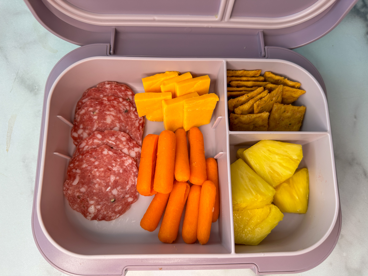 A kids lunch box with sliced salami, carrot sticks, pineapple chunks, healthy crackers, cheese and cheese squares.