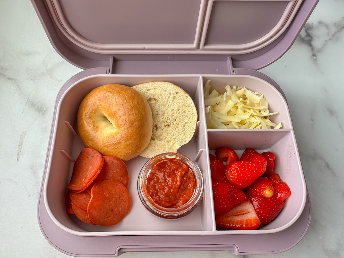 Kids lunchbox with a mini bagel, turkey pepperonis, a small jar of pizza sauce, shredded mozzarella, and cut up strawberries