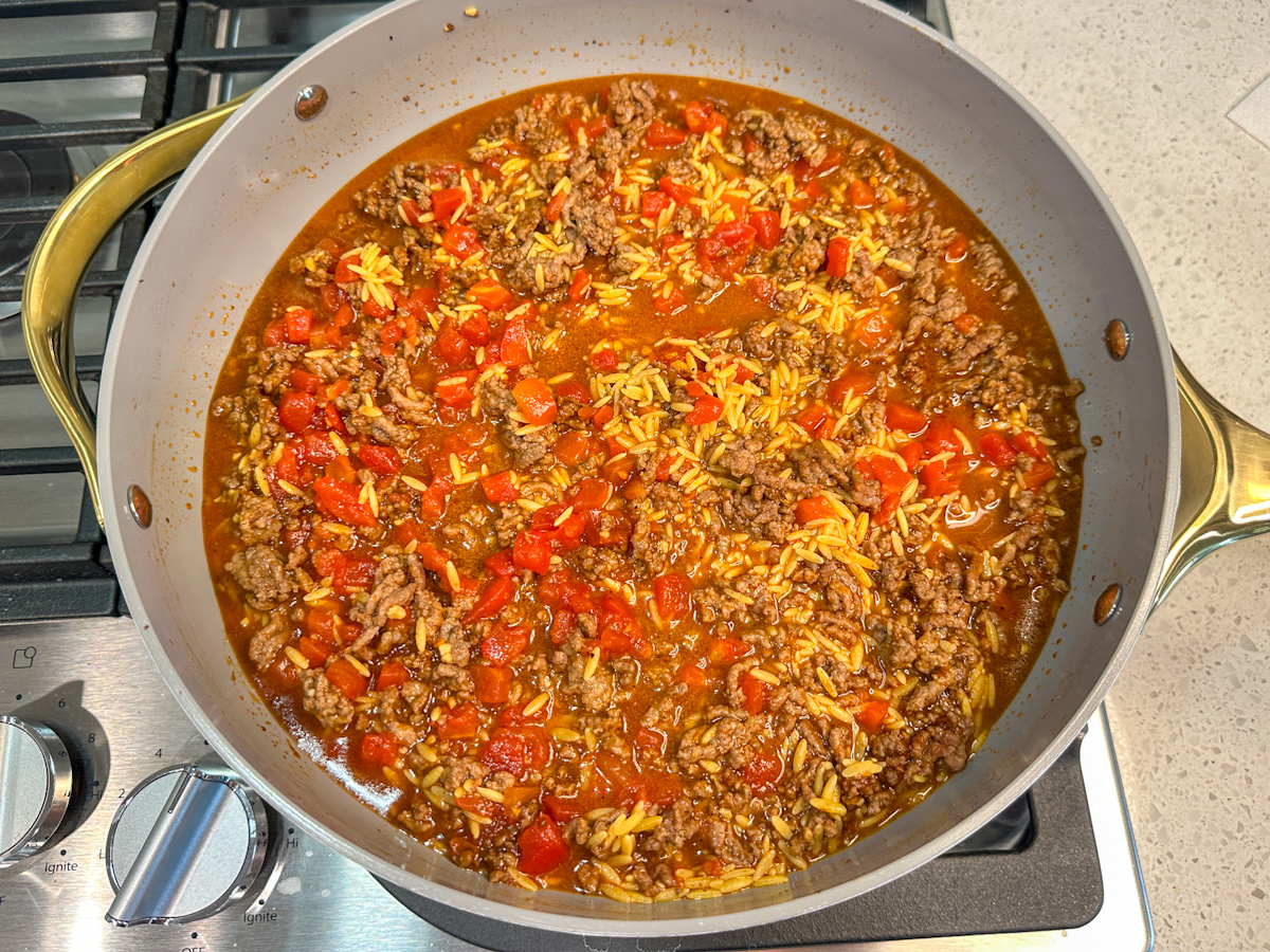 Large skillet with cooked ground beef, orzo, diced tomato, and liquid.